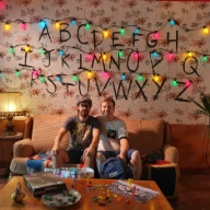 Two beautiful guys sitting in a couch which is on the set of Stranger Things with Christmas alphabet lights
